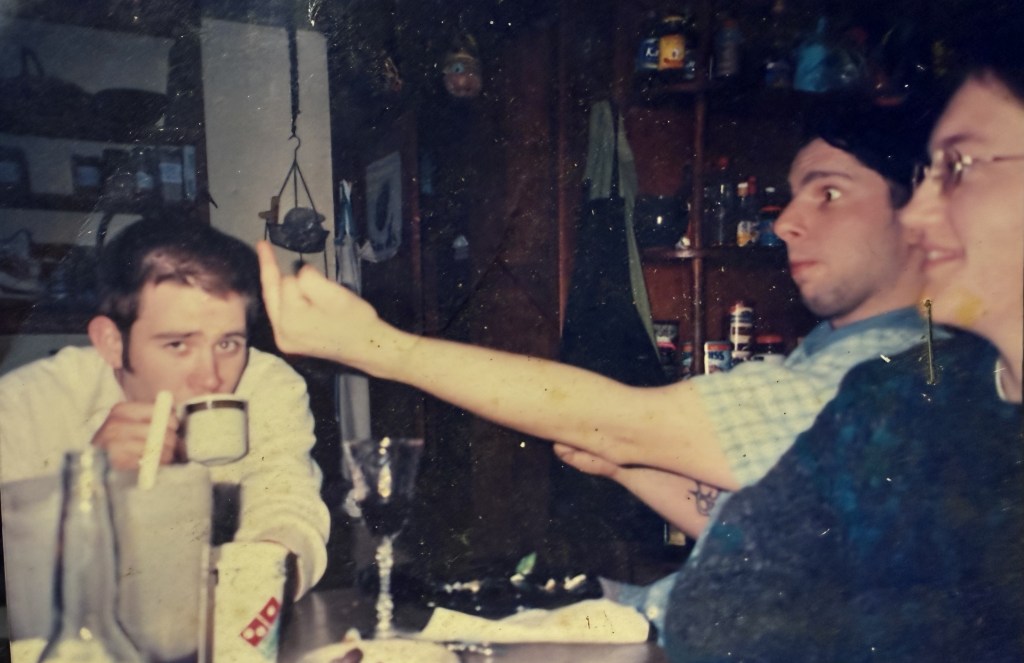 Three men at the dinner table by candlelight, one reaching across the frame, mugs and papers on the table.