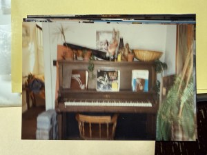 An upright piano with potted plants and books arranged on top, a wooden chair pulled up to it.