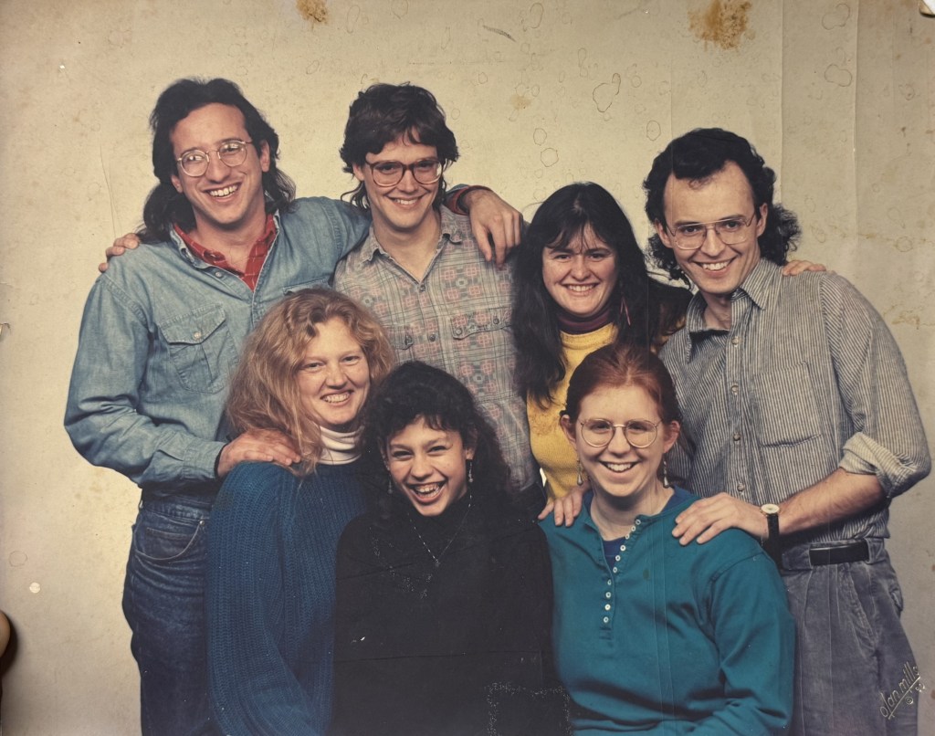 A posed group portrait of seven people against a mottled studio backdrop, arms around each other.