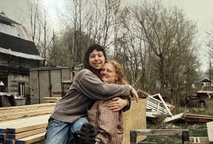 Two people embracing beside a stack of new lumber and a pile of demolition debris, an outbuilding behind them.
